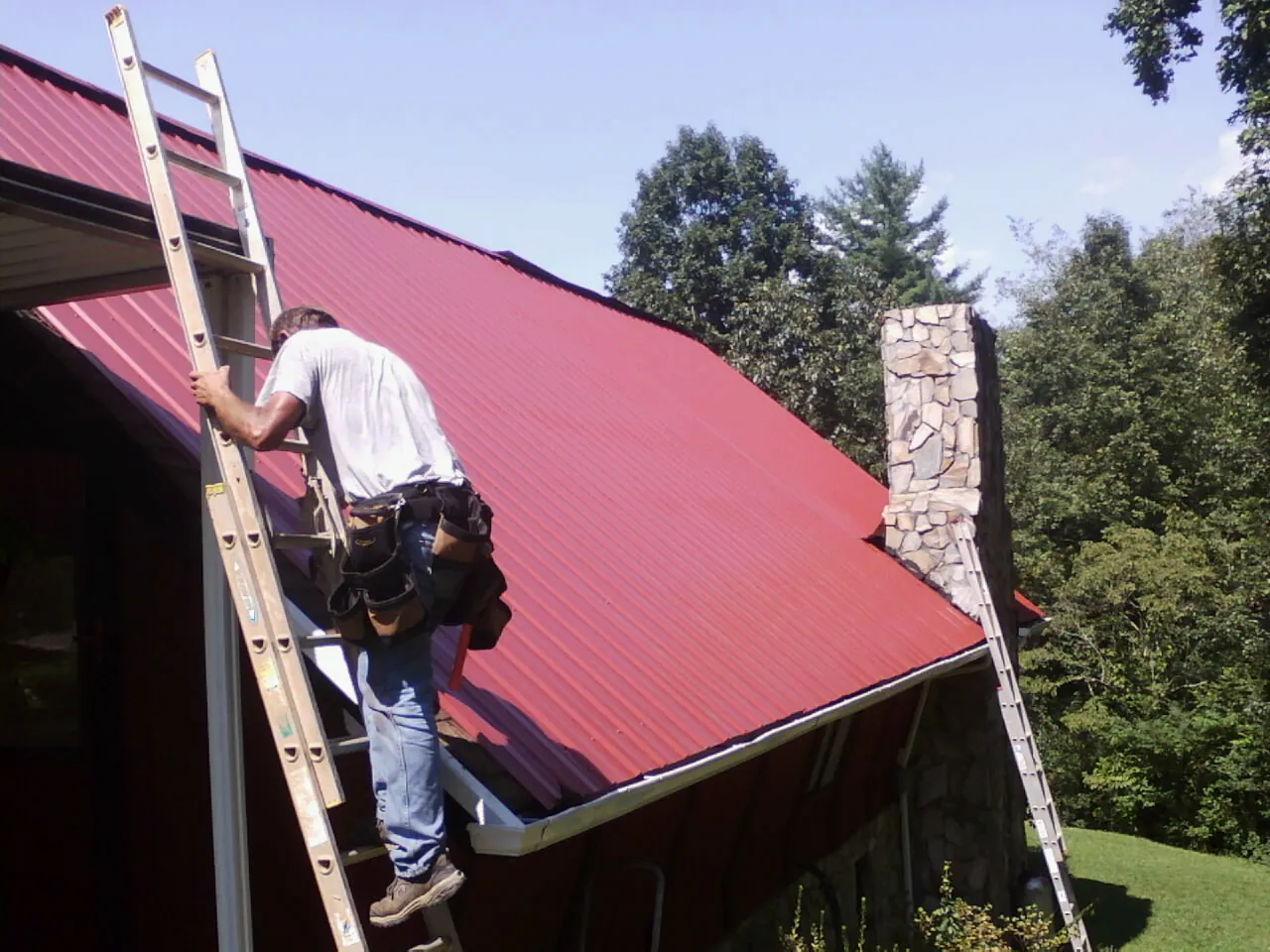 Roofer on a ladder beside a red metal roof and stone chimney — Soffit & Fascia Repair by Roofing Brothers in Cyril